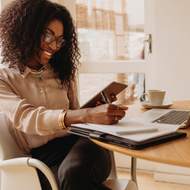 Woman with curly hair and glasses smiling while using a tablet at cafe table with notebook and coffee.