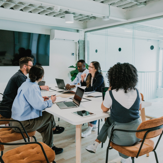 Diverse team collaborating at conference table with laptops in modern office space with glass partitions.