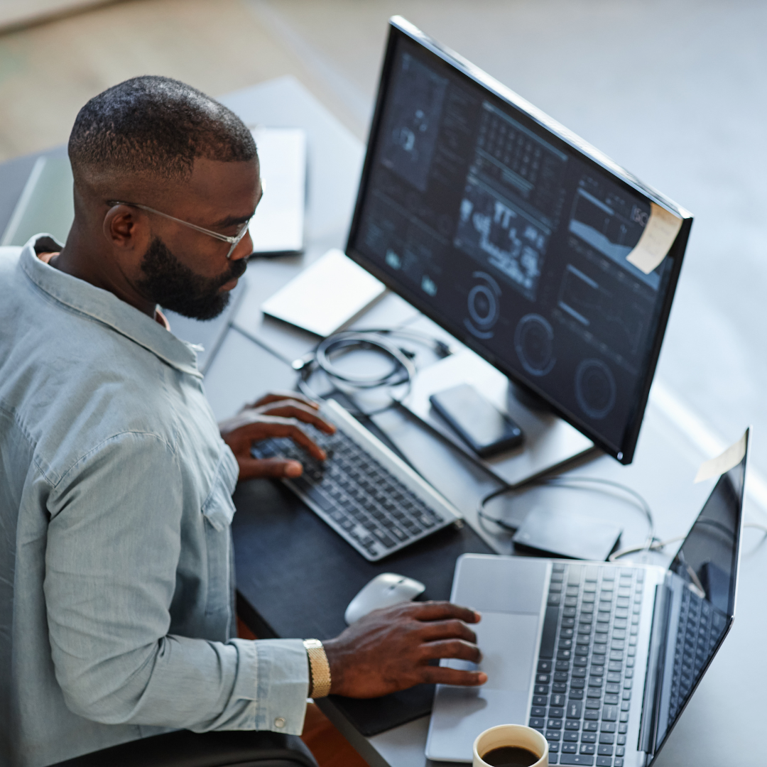 Man wearing a white jacket is working at a computer desk with a desktop computer and laptop.
